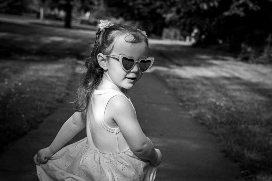 A girl in heart-shaped sunglasses on a tree-lined path, in black and white